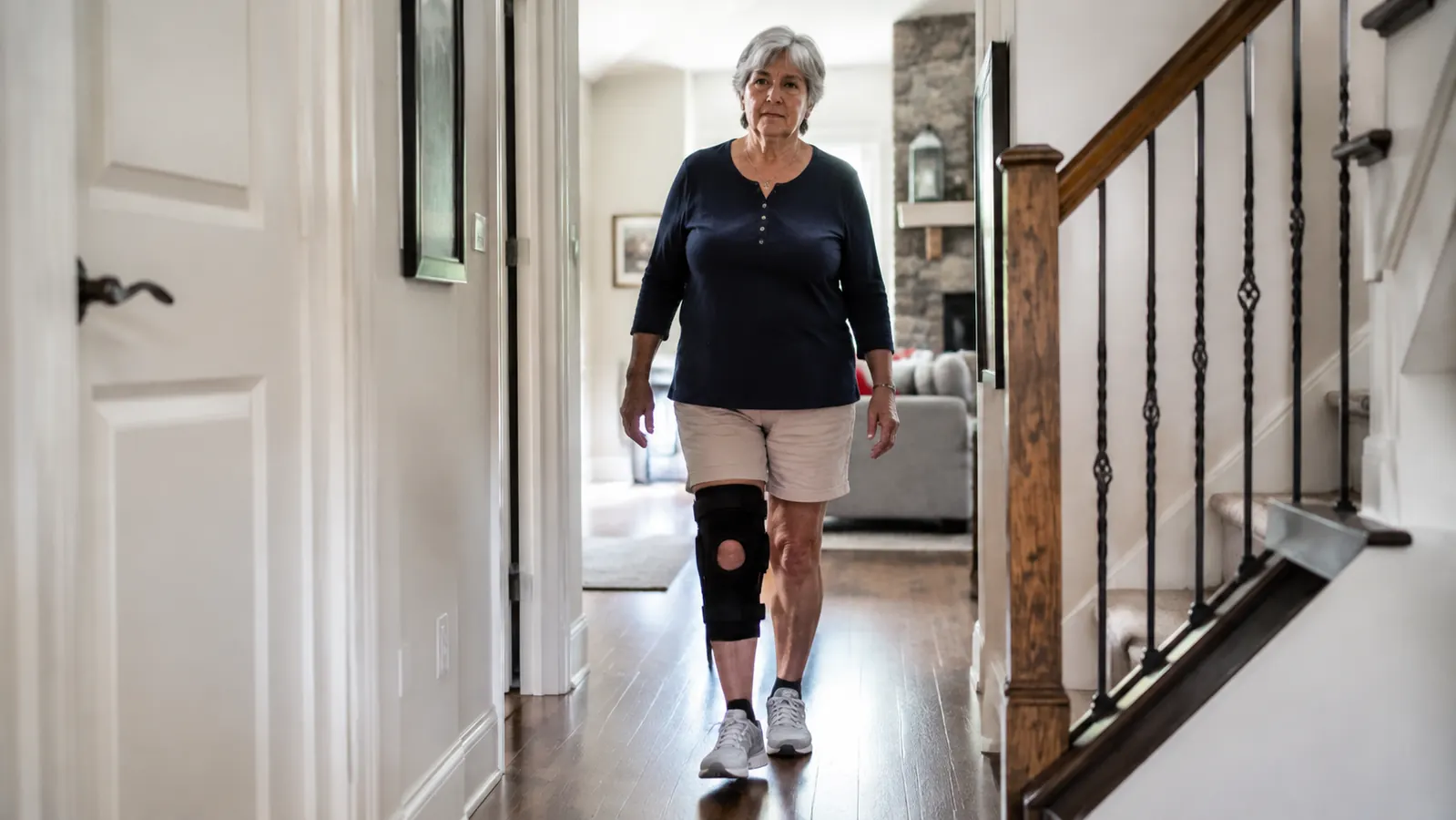 A woman in her sixties walking down a bright hallway in a home, a knee brace visible on her left leg, everyday domestic setting
